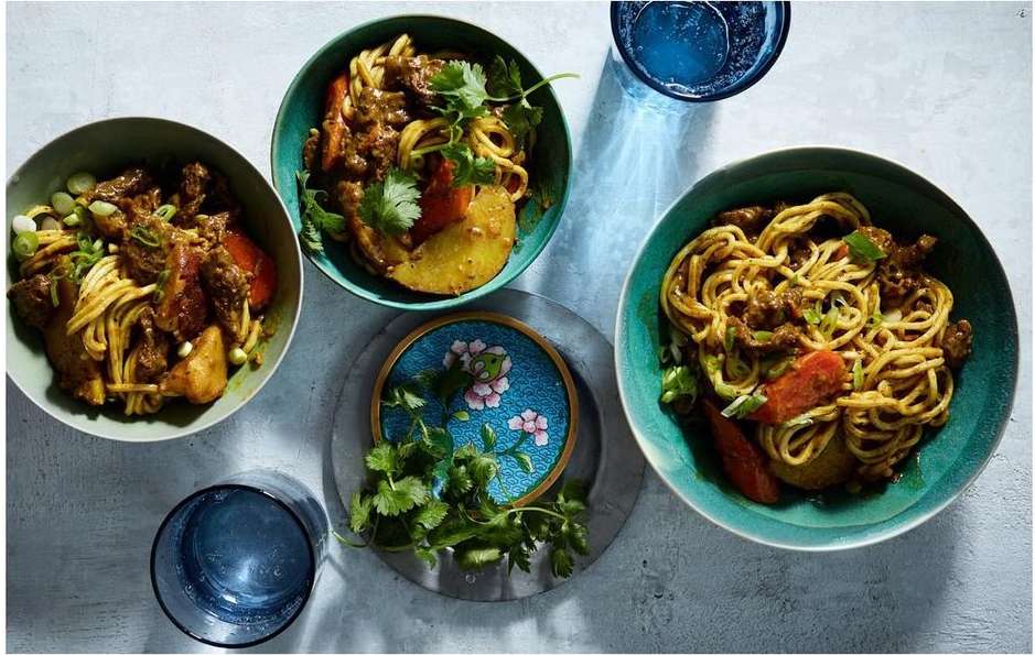 Three bowls of Asian noodles with beef, carrots, potatoes, and green onions, alongside blue-tinted water glasses on a grey surface.