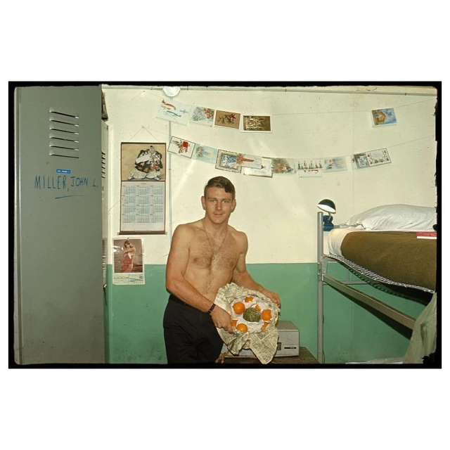 Man in barracks holds fruit basket with bunk beds and locker. Candid photo of adult male indoors with vintage military style decor.