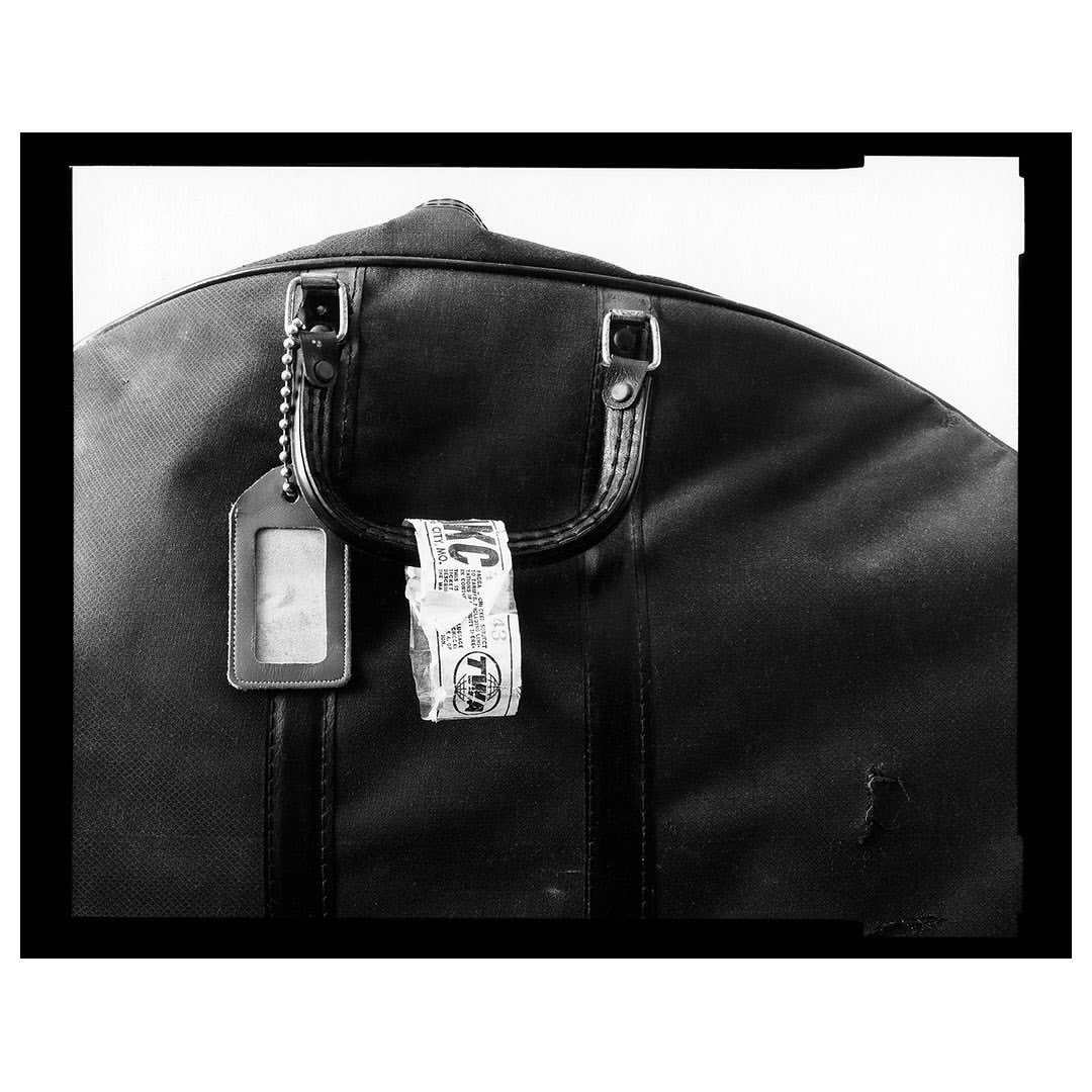 Black garment bag with a luggage tag and baggage claim tag against a white background, with a black border creating a studio-like feel.