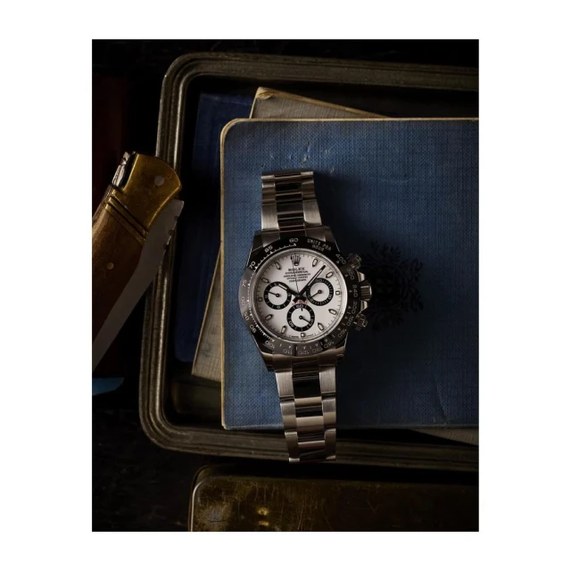 Wristwatch with silver stainless steel band and white dial face on blue books with silver tray and brown pocket knife, product photography.