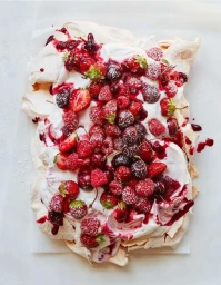 Top-down view of a pavlova dessert topped with whipped cream, red berries, and fruit coulis on white parchment paper