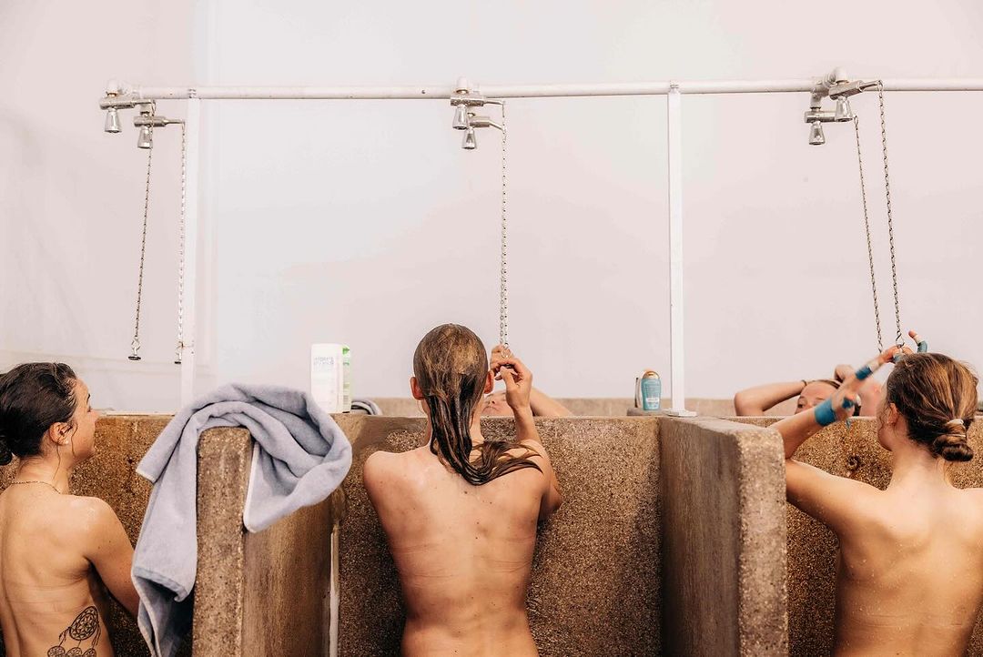 Three women standing in individual shower stalls with towel and toiletries, shot at eye-level in bright, natural light.