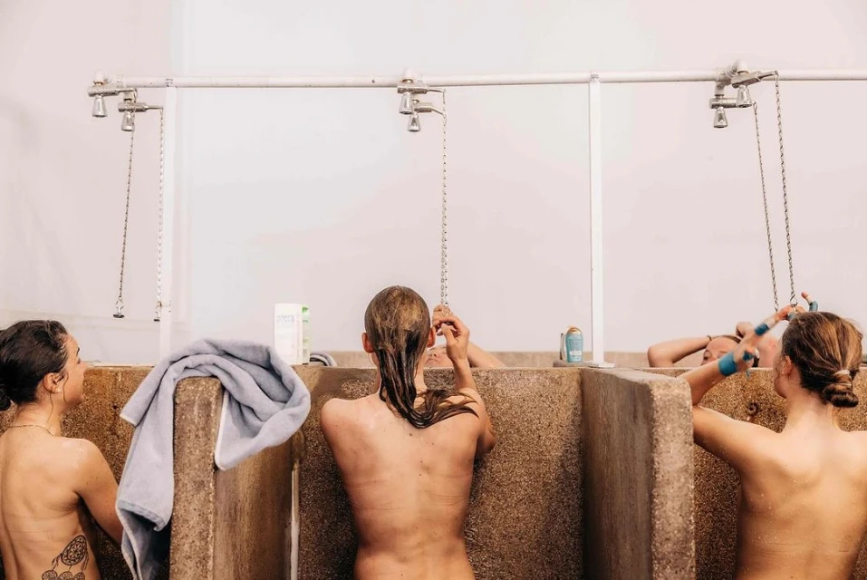 Three women standing in individual shower stalls with towel and toiletries, shot at eye-level in bright, natural light.
