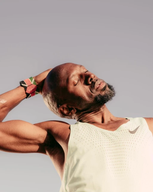 Bald African-American athlete stretches his neck muscles outside in sportswear against a grey background, wearing a beaded bracelet.