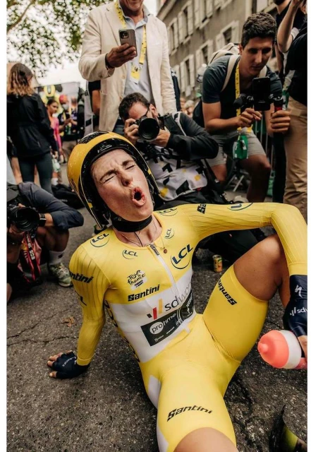 Female cyclist in yellow uniform sitting on the ground, looking exhausted after race, surrounded by photographers and onlookers.