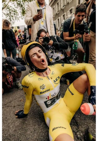 Female cyclist in yellow uniform sitting on the ground, looking exhausted after race, surrounded by photographers and onlookers.