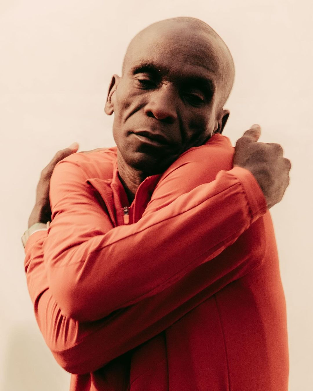 Adult male with a bald head wearing a red long sleeve shirt with a zipper, hugging himself in front of a plain background.