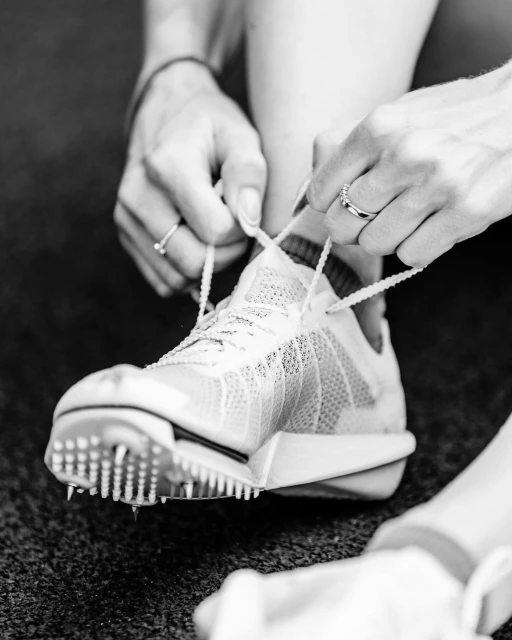 Close-up black and white shot of a person tying the laces of a spiked athletic shoe on a dark surface, illuminated by soft lighting.