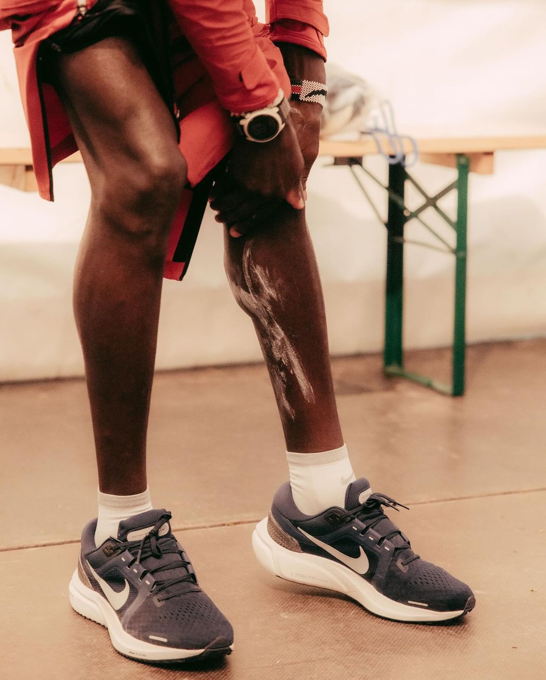 Close-up of an adult male applying lotion to his leg, wearing running shoes and a smart watch, standing indoors by a table.