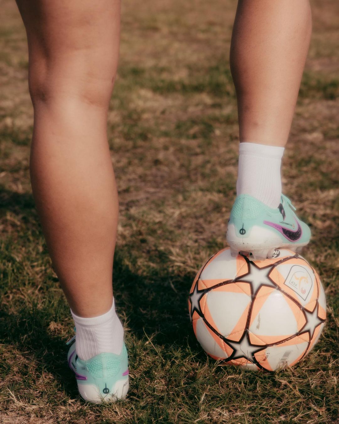 Athlete's feet and soccer ball on grass. Soccer shoes in white and teal, white socks, and a white ball with orange stars visible.