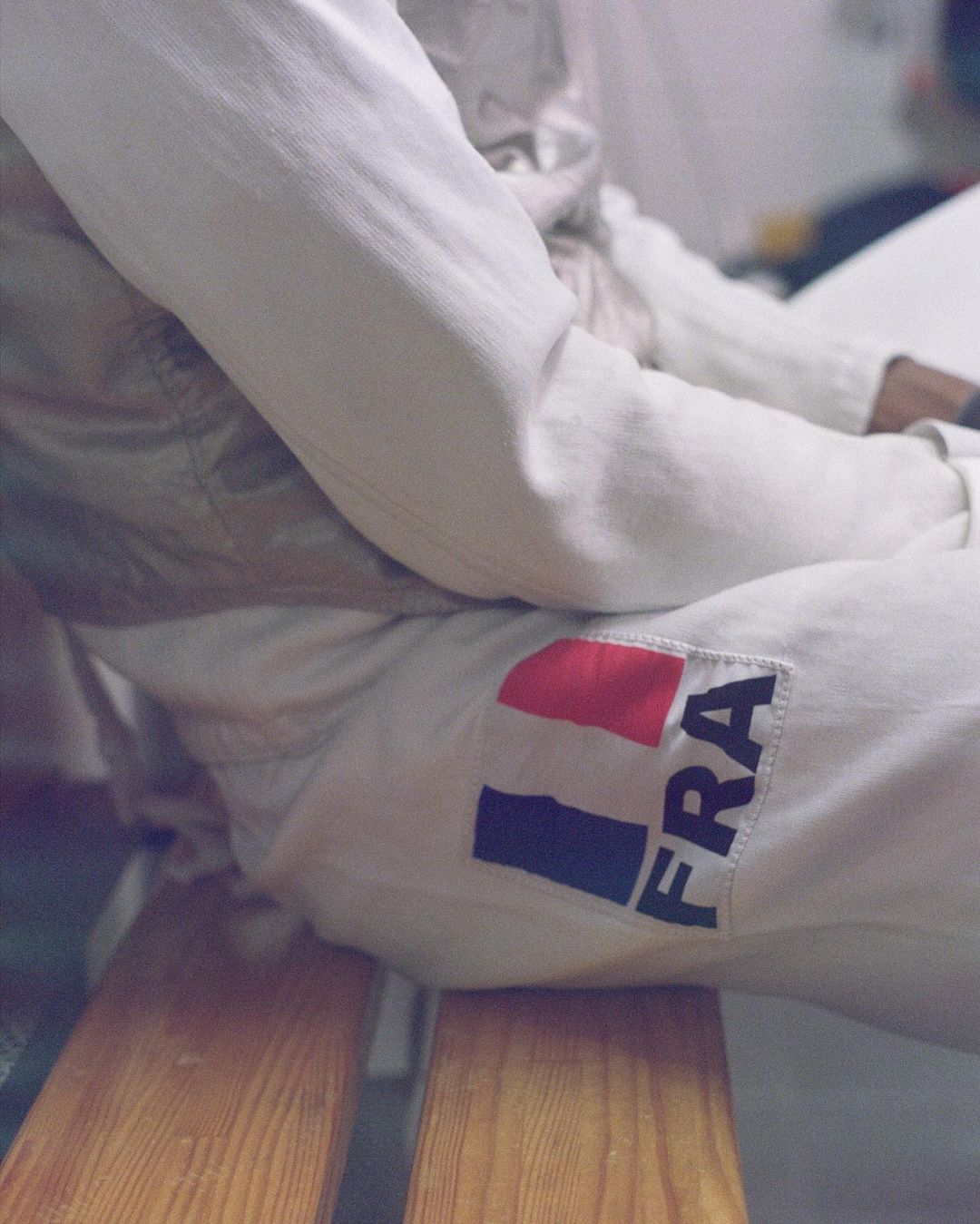 Close-up of a person wearing a white fencing uniform with a French flag patch, seated on a wooden bench indoors.