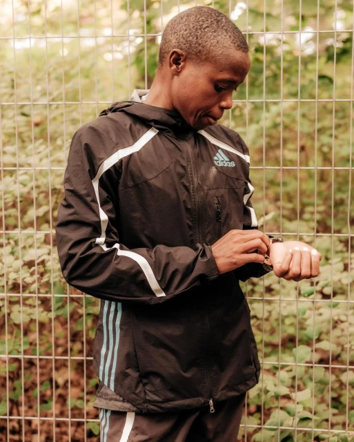 Adult in an Adidas jacket checks a smartwatch in front of a metal fence with green foliage, captured in a medium shot with natural lighting.