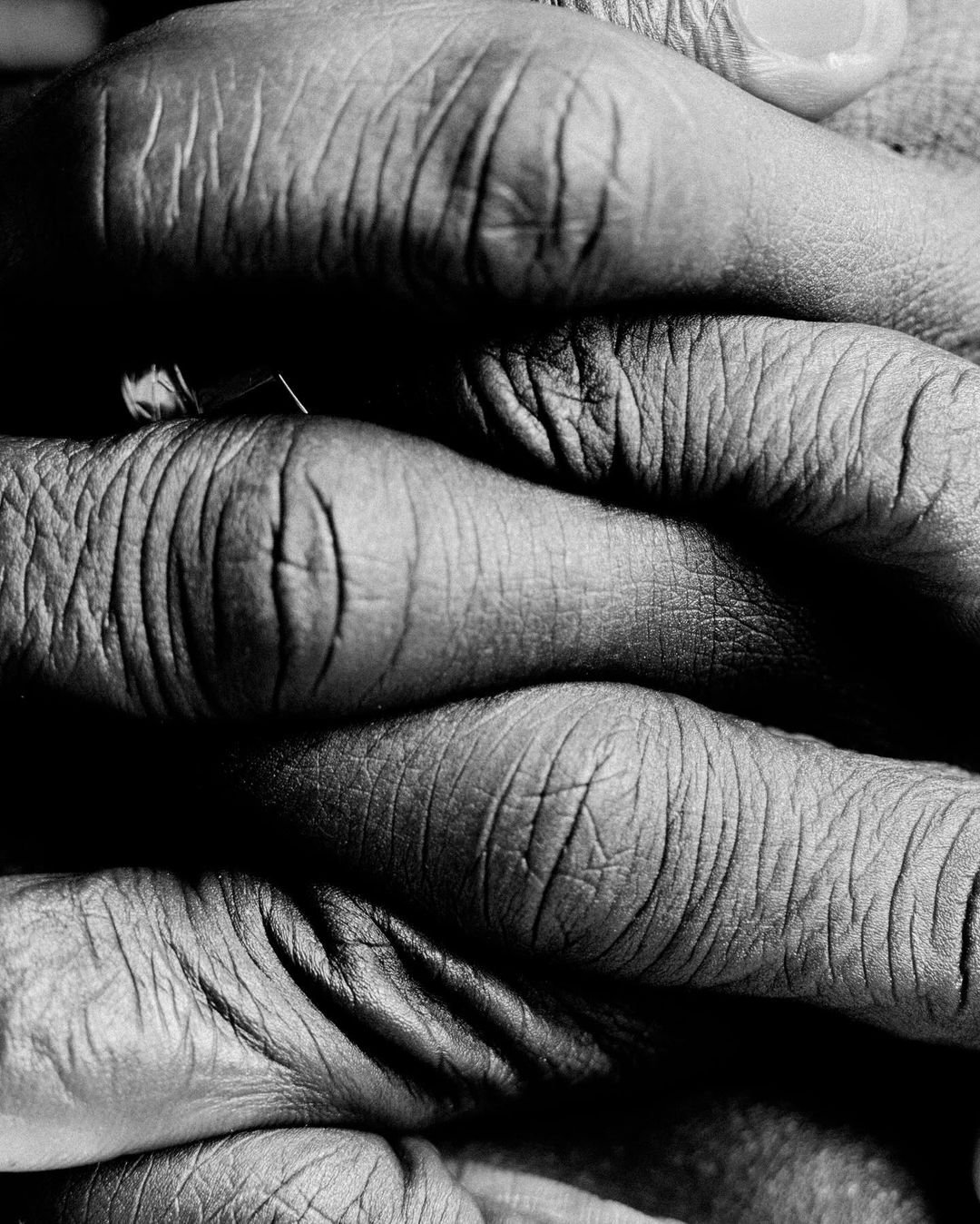 Monochrome image showing the close-up of an interlaced and stacked set of human hands, displaying skin texture and contrasting shadows.