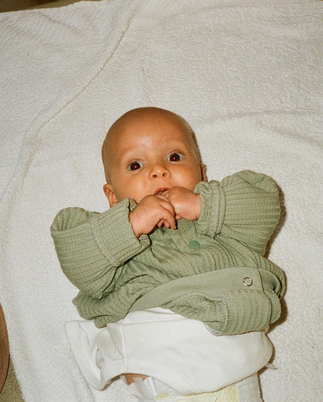 Close-up of a baby lying on their back on a white towel, wearing a light green, button-up shirt, and a white diaper.