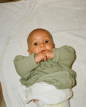 Close-up of a baby lying on their back on a white towel, wearing a light green, button-up shirt, and a white diaper.