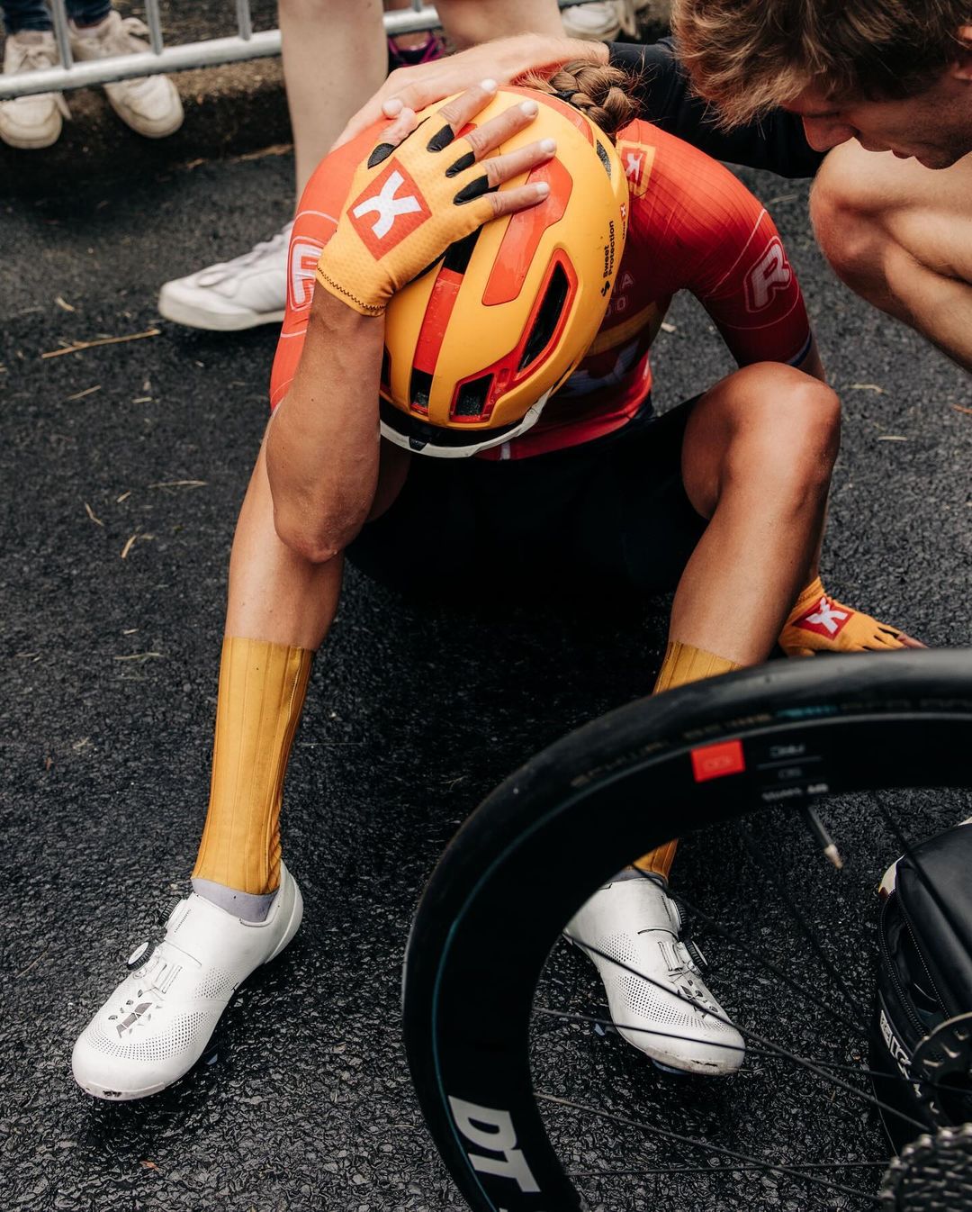 Cyclist in red and yellow with helmet sits on asphalt with bike wheel visible; support staff around him; daytime, outdoor sport accident.