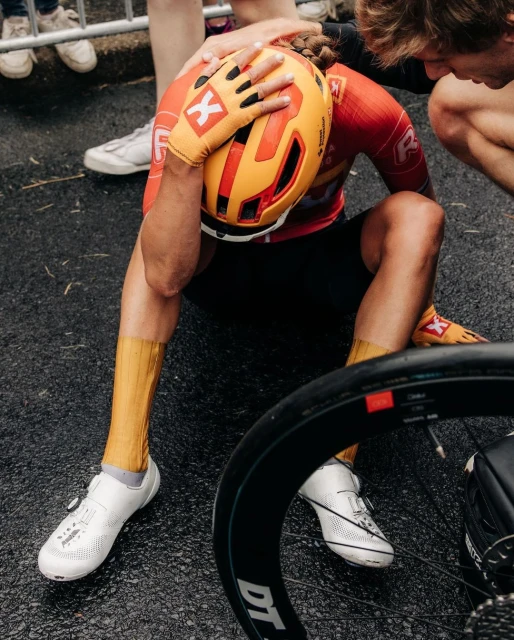 Cyclist in red and yellow with helmet sits on asphalt with bike wheel visible; support staff around him; daytime, outdoor sport accident.