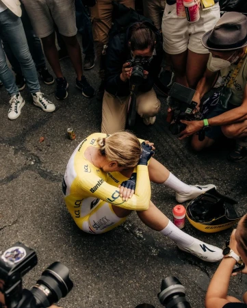 Professional female cyclist in yellow uniform sitting on the road, surrounded by photographers at an outdoor sporting event.