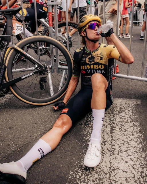 Professional male cyclist drinks water after a race. He wears a yellow-and-black uniform and helmet, sitting near his bicycle on the road.
