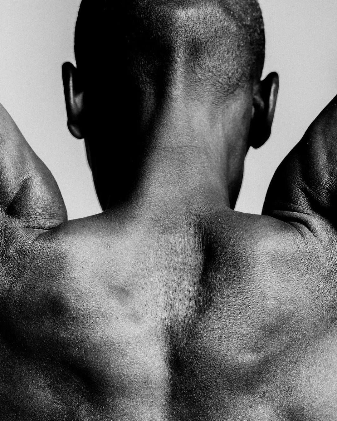 Close-up black and white photo of the bare back and neck of a dark-skinned adult male with short hair, arms raised, against a plain light background.