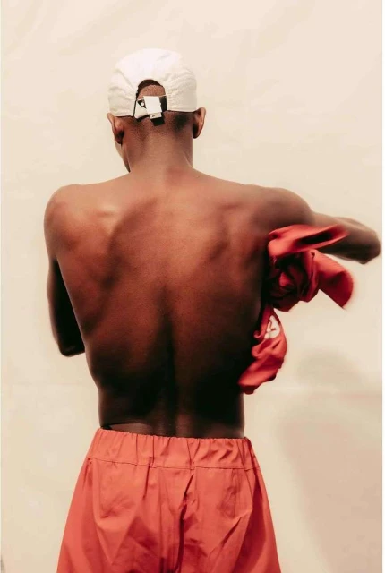 Adult man with bare back standing with white cap and red shorts holding a red cloth on his shoulder indoors in front of wall.