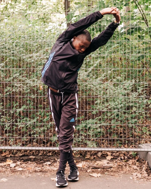 Young Black male in black Adidas tracksuit stretches arms overhead in outdoor setting with fence and foliage backdrop, sportswear fitness.