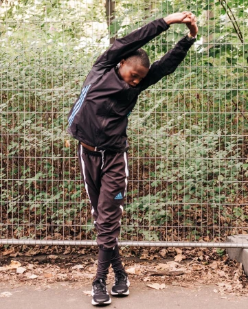 Young Black male in black Adidas tracksuit stretches arms overhead in outdoor setting with fence and foliage backdrop, sportswear fitness.