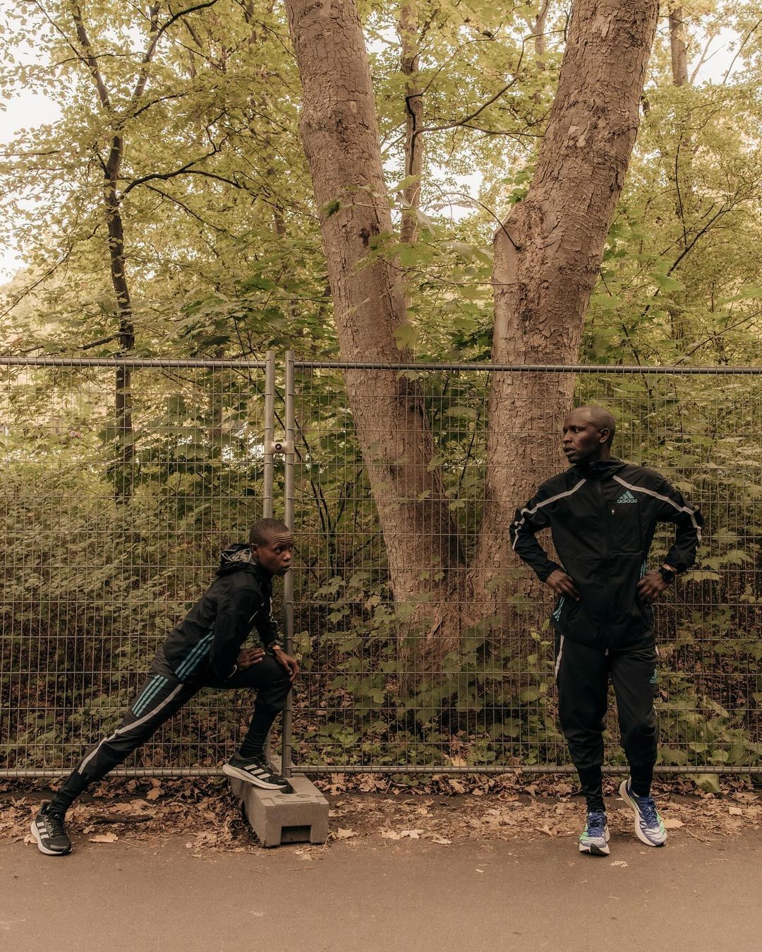 Two adult males in athletic wear stretch near a metal fence in a wooded area with soft, natural lighting, showcasing fitness and outdoor activity.