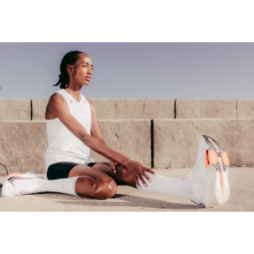 Athlete stretching legs outdoors while seated on concrete ground wearing white sports apparel and sneakers.