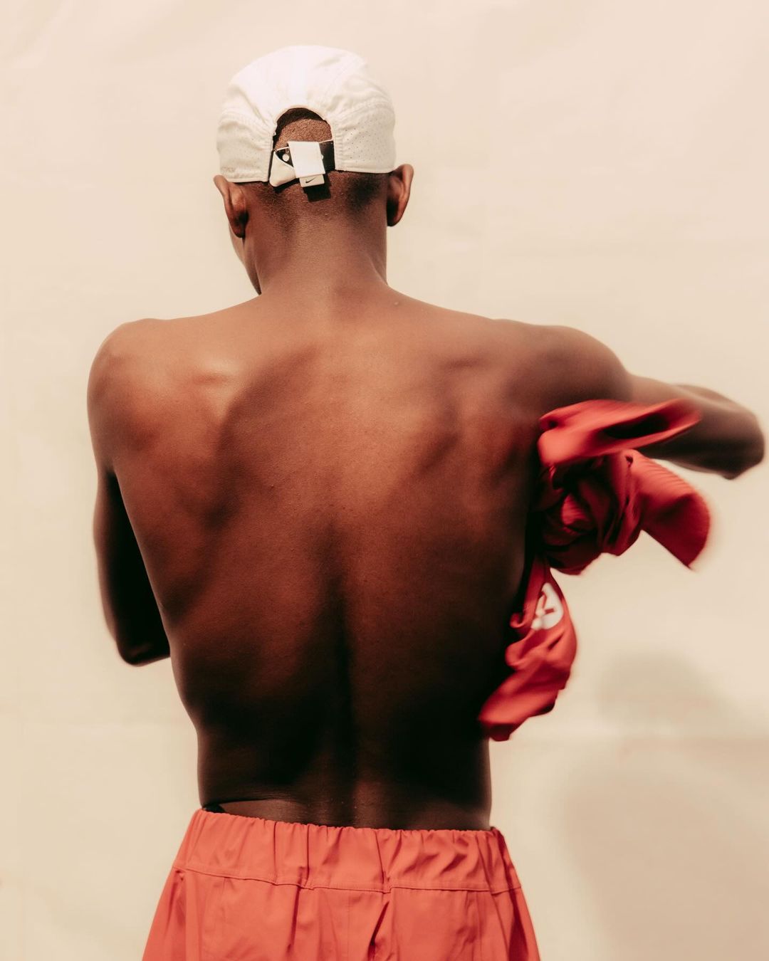 Back view of a shirtless Black man wearing a white baseball cap and orange shorts, holding an orange cloth indoors.