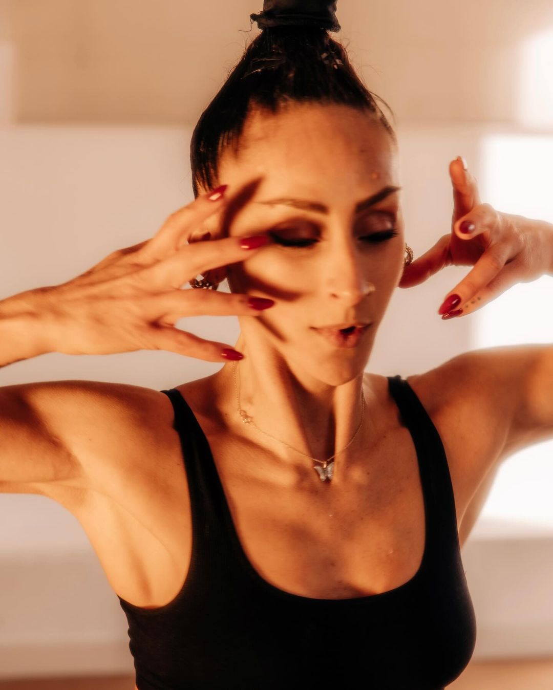 Portrait of adult woman with dark hair in a bun, hands near her face, wearing a black tank top, a butterfly necklace, and red nail polish.