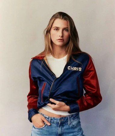 Woman wearing blue and red satin jacket with the name Chris on the chest, a white t-shirt, and blue jeans against a neutral background.
