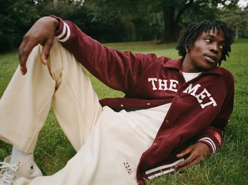 An adult man with dreadlocks lounges on green grass wearing a burgundy letterman jacket with white lettering and tan pants.