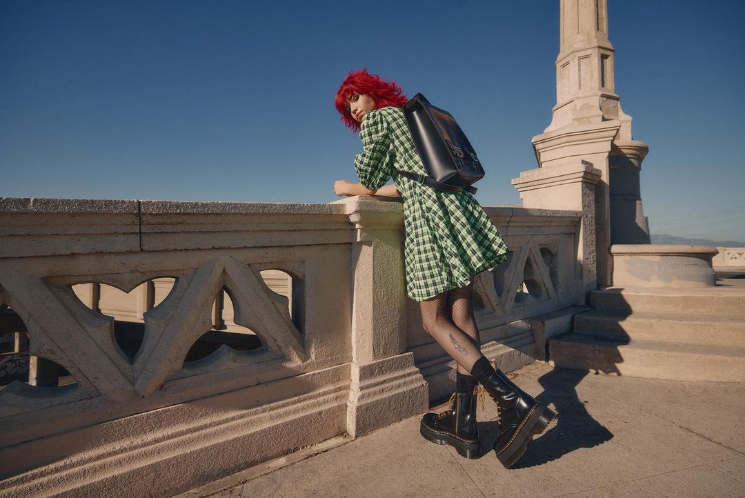 Woman with red hair and backpack wearing a green plaid dress and black boots standing on a stone ledge against a blue sky.