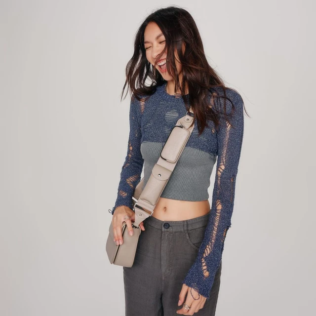 Smiling Asian woman in a ripped blue long sleeve crop top, gray ribbed tank top, and gray pants with a crossbody bag in a studio.