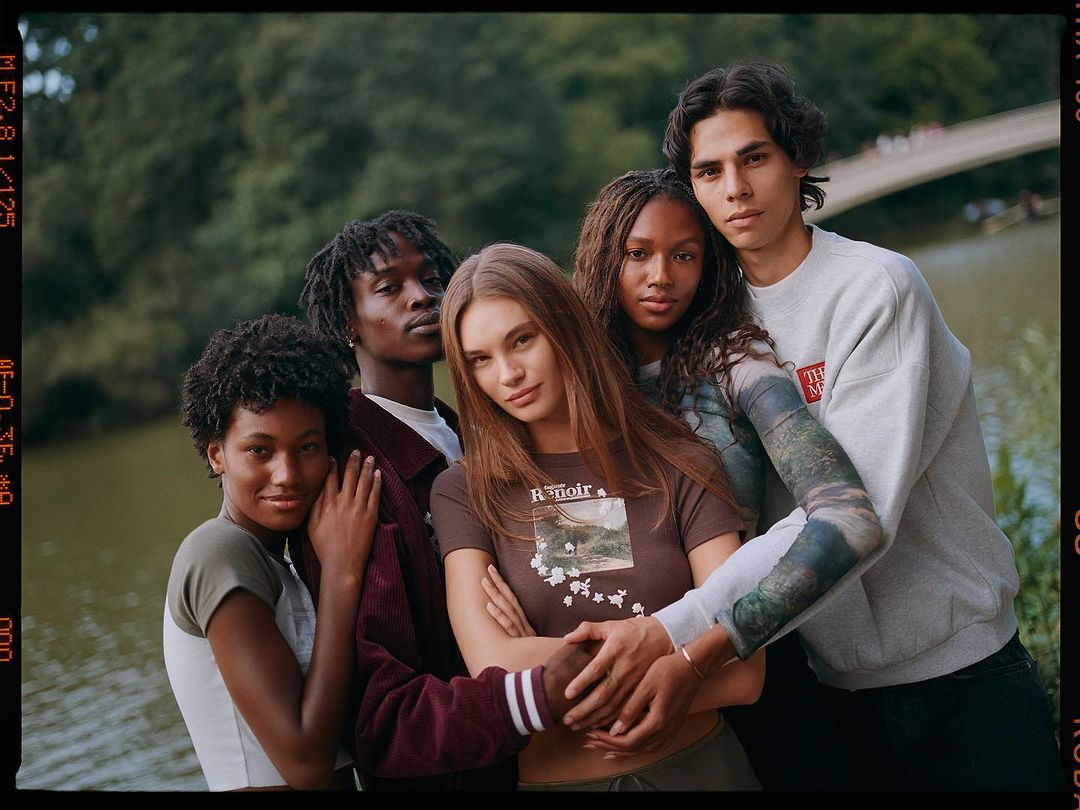 Group of five young adult diverse people are standing outside in front of water with trees and a bridge, fashion shot.