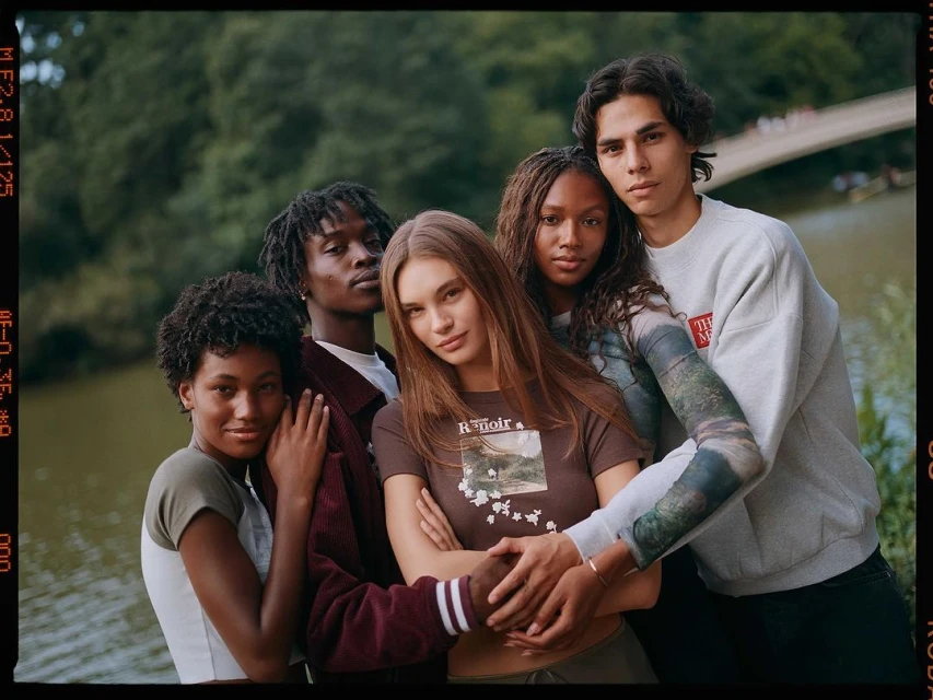 Group of five young adult diverse people are standing outside in front of water with trees and a bridge, fashion shot.