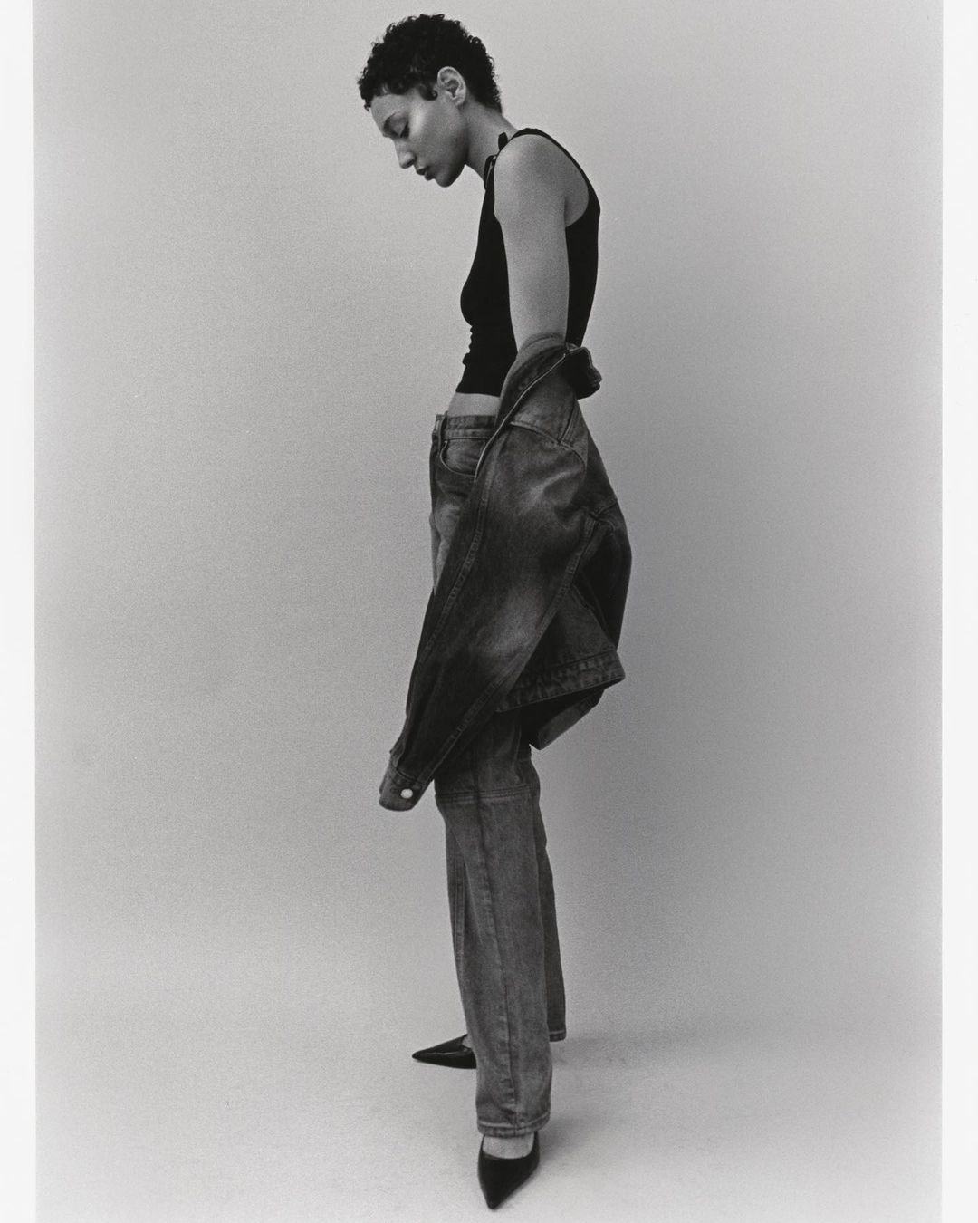 Model with short dark hair wearing a black tank top and denim jeans against a plain studio background, photographed by Jon Stars.