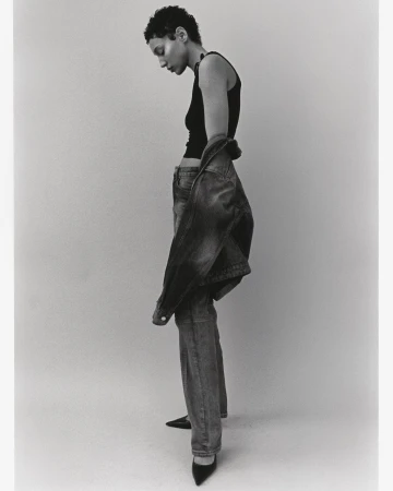 Model with short dark hair wearing a black tank top and denim jeans against a plain studio background, photographed by Jon Stars.