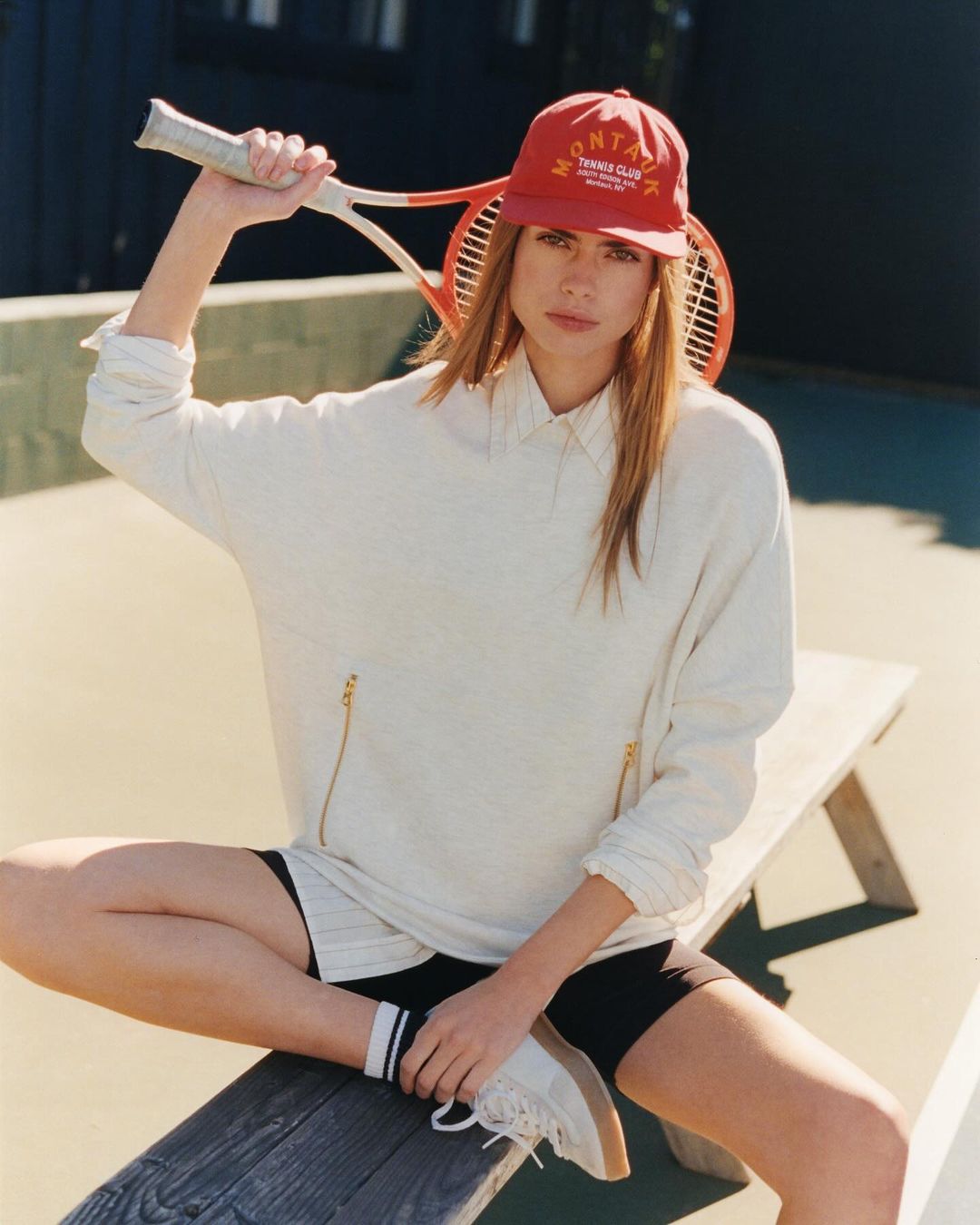 Woman in red cap and sweatshirt holds tennis racket, sitting on a bench at an outdoor tennis court on a sunny day.
