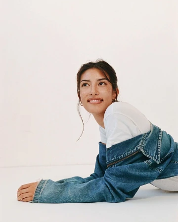 Woman in denim jacket over white t-shirt lying on white studio floor looking up