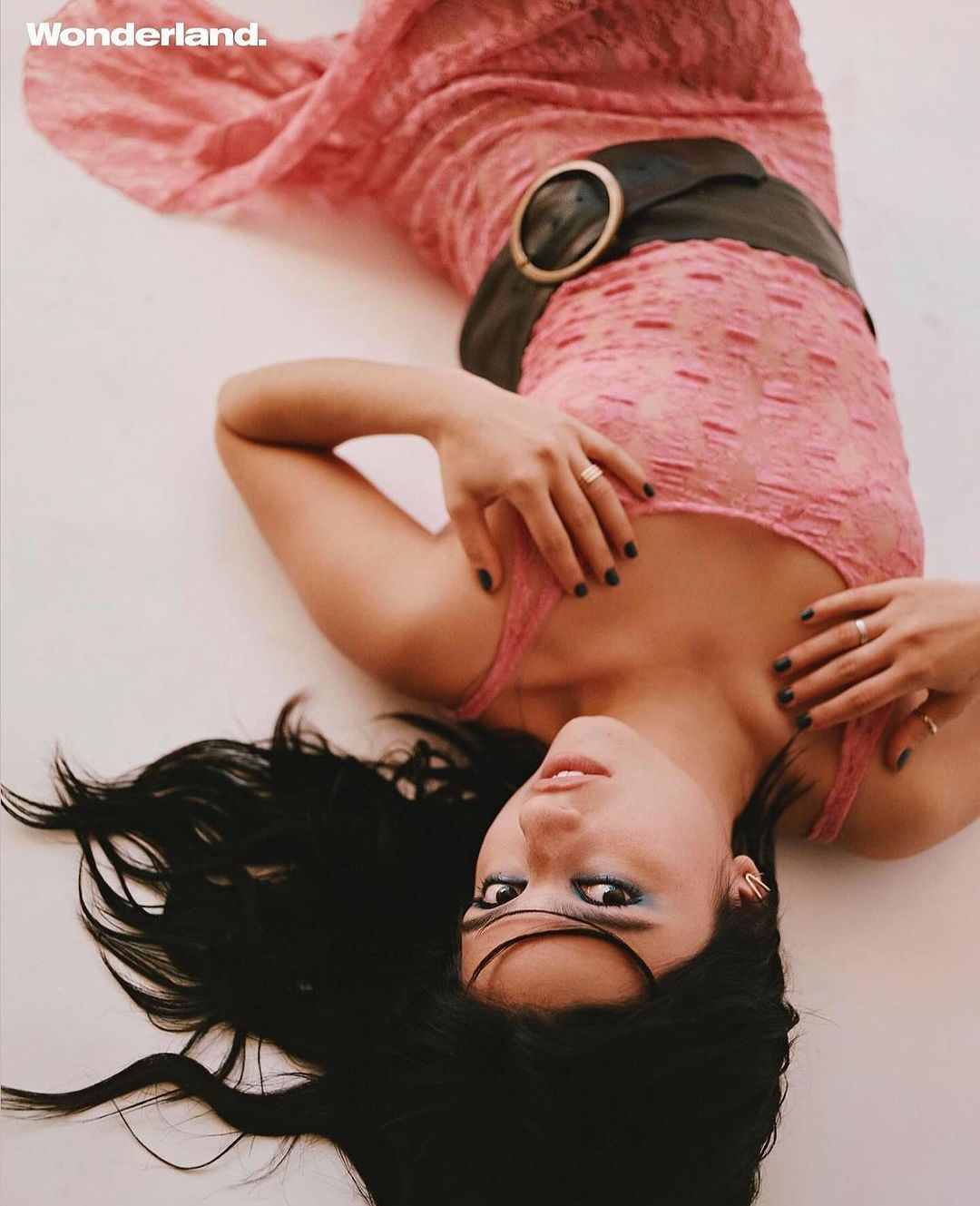 Woman in pink lace dress with a large belt, lying down on a white surface with dark hair and dark nail polish, looking at the camera.