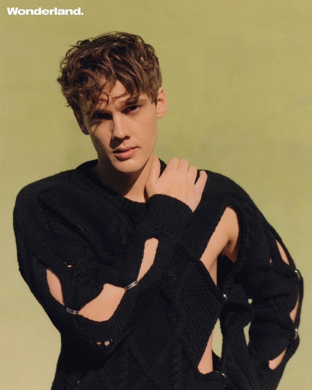 Close-up shot of a fair-skinned young man in a black, cut-out knit sweater against a light green background for Wonderland magazine.