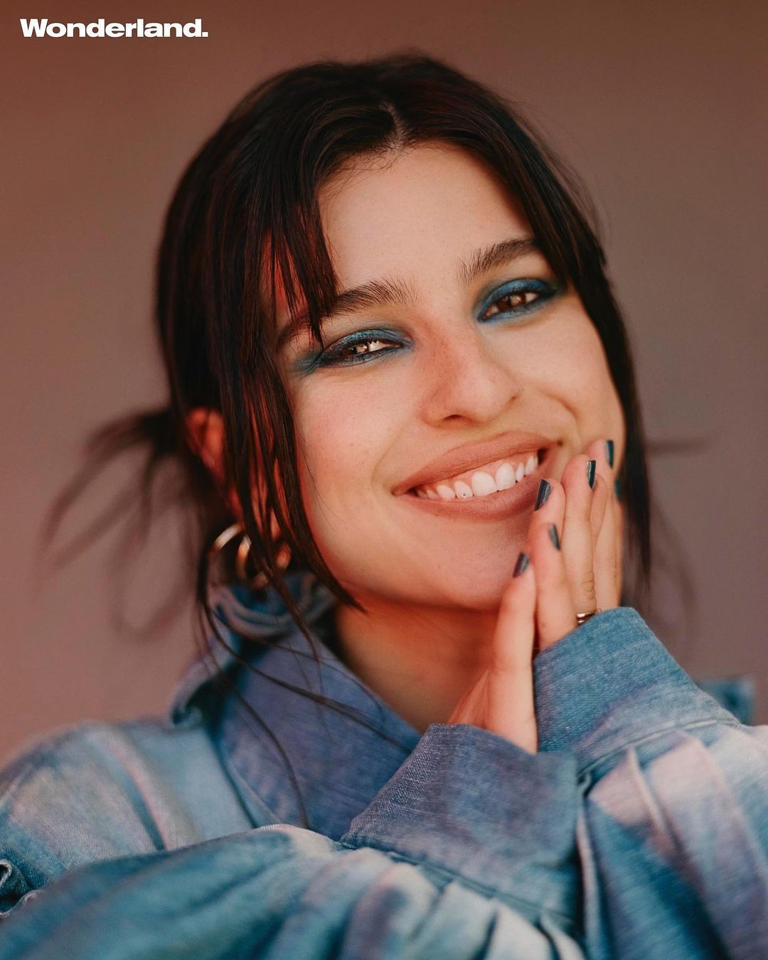 Close-up portrait of a smiling woman with dark hair and blue eyeshadow, wearing a denim top against a warm-toned background.