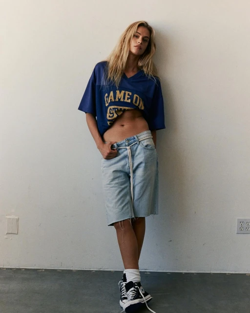 Blonde woman in a blue football jersey and denim shorts leans against a white wall in a fashion style full portrait.