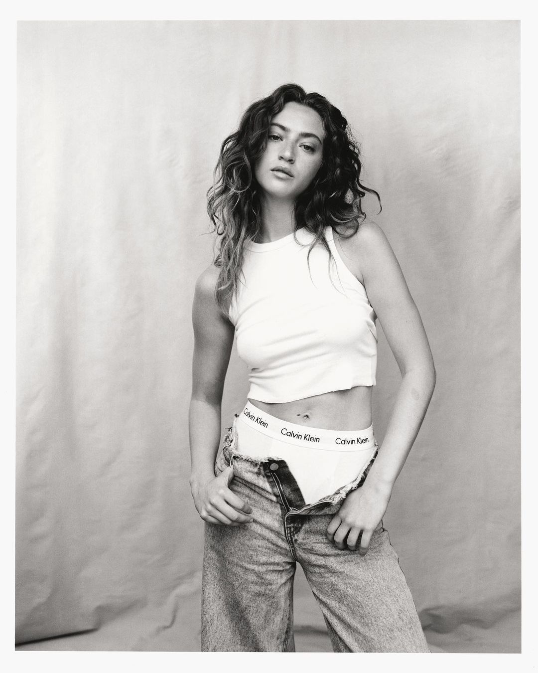 Young woman poses in a white tank top and unbuttoned jeans, exposing branded underwear against a neutral backdrop, casting a casual mood.