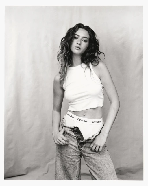 Young woman poses in a white tank top and unbuttoned jeans, exposing branded underwear against a neutral backdrop, casting a casual mood.