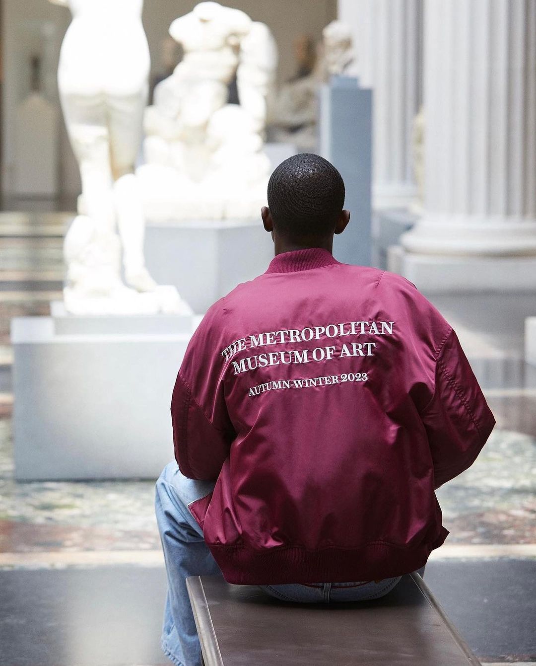 An African-American adult male sitting on a bench at the Metropolitan Museum of Art, wearing a maroon jacket with white text, medium shot.