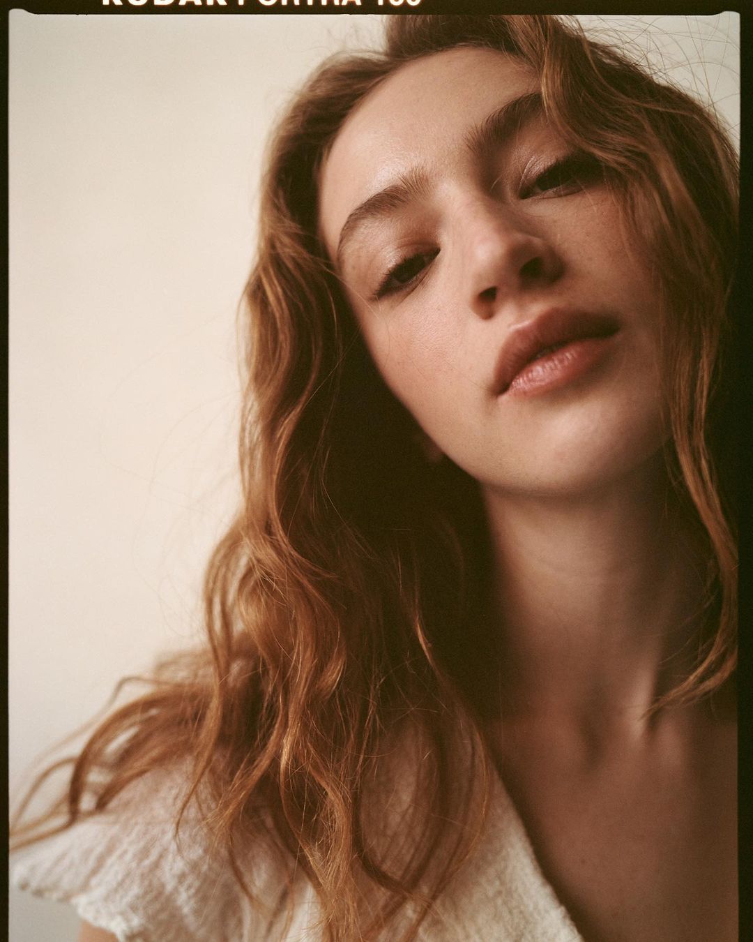 Portrait of a young woman with reddish-brown wavy hair, fair skin, and neutral expression in a close-up shot, soft lighting.