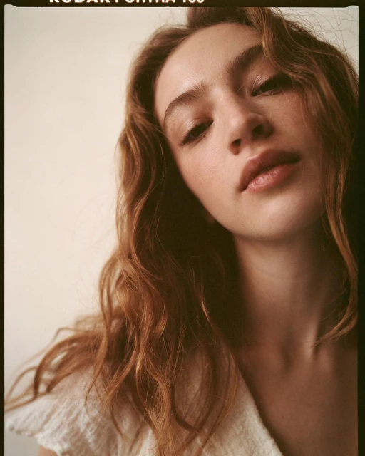 Portrait of a young woman with reddish-brown wavy hair, fair skin, and neutral expression in a close-up shot, soft lighting.